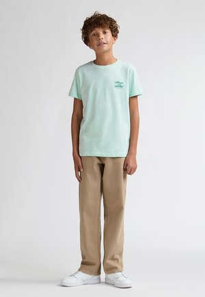 T-shirt en coton vert menthe à manches courtes et col rond, avec un petit logo imprimé. Associé à un pantalon beige et des baskets blanches.