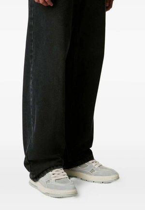 Persona che indossa pantaloni neri a gamba larga e sneaker grigio chiaro con lacci bianchi, in piedi su una superficie bianca.