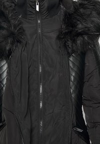 Chaqueta acolchada negra con capucha de pelo sintético, detalles de cuero acolchado y cremallera central en el frente. Textura suave con puños acanalados.