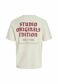 Cremefarvet T-shirt med fed rød tekst "Studio Originals Edition" og mindre beskrivende sætninger på bagsiden.