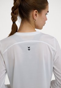 Weißes, leichtes Sportshirt aus strukturiertem Stoff, mit einem schwarzen Chevron-Logo auf dem Rücken und einem runden Halsausschnitt.