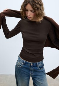 Maglia marrone a maniche lunghe realizzata in tessuto morbido, abbinata a jeans in denim azzurro chiaro con vestibilità leggermente ampia e aree scolorite.