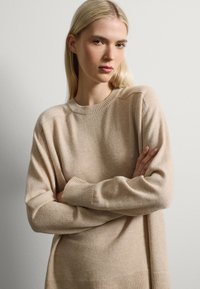 Beige gebreide trui met een ribbelpatroon en ronde halslijn en manchetten. Heeft een licht oversized pasvorm, zachte textuur en subtiele kleurvariaties.