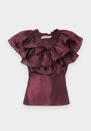 Femme portant un haut sans manches bordeaux à volants et une jupe asymétrique fleurie aux tons roses et jaunes, assortis à des talons nus avec bride à la cheville.