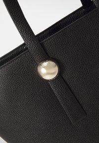 Sac fourre-tout en cuir noir avec une surface texturée, comportant un bouton arrondi en argent avec le nom de la marque gravé. Longues anses plates.