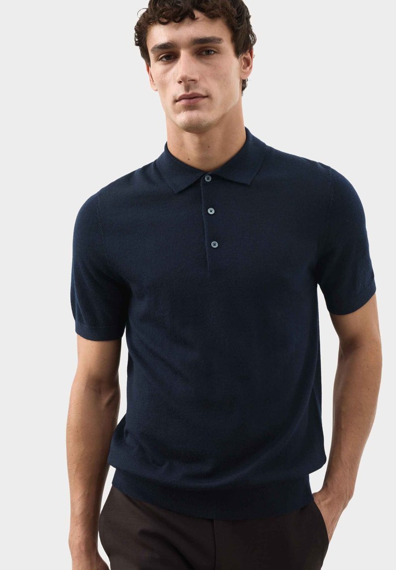 Giovane uomo che indossa una polo blu navy a maniche corte con tre bottoni, posando davanti a uno sfondo chiaro e uniforme.