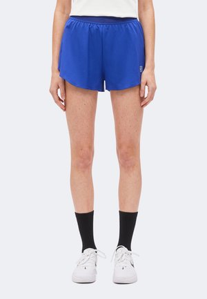 Personne portant un short de sport bleu vif, des chaussettes noires montantes et des baskets blanches, debout les bras détendus le long du corps.