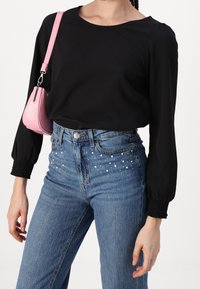 Haut noir à manches longues avec des poignets froncés, associé à un jean en denim bleu avec des accents décoratifs de perles blanches le long de la taille. Sac à main rose.