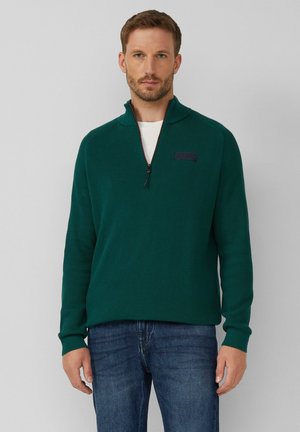 Grüner, strukturierten Strickpullover mit einem Viertelreißverschluss und einem dunklen Etikett auf der Brust. Verfügt über lange Ärmel und einen lockeren Schnitt.