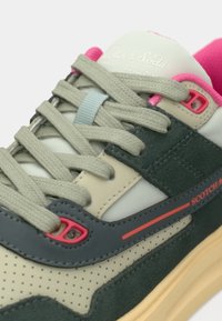 Gros plan d'une basket beige et vert foncé avec des lacets gris, des accents roses, des détails perforés et le nom de la marque sur le côté.