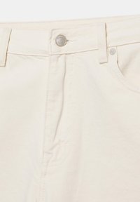 Gros plan sur la ceinture d’un jean en denim blanc cassé avec un bouton en métal et une boucle de ceinture au-dessus de la poche avant.