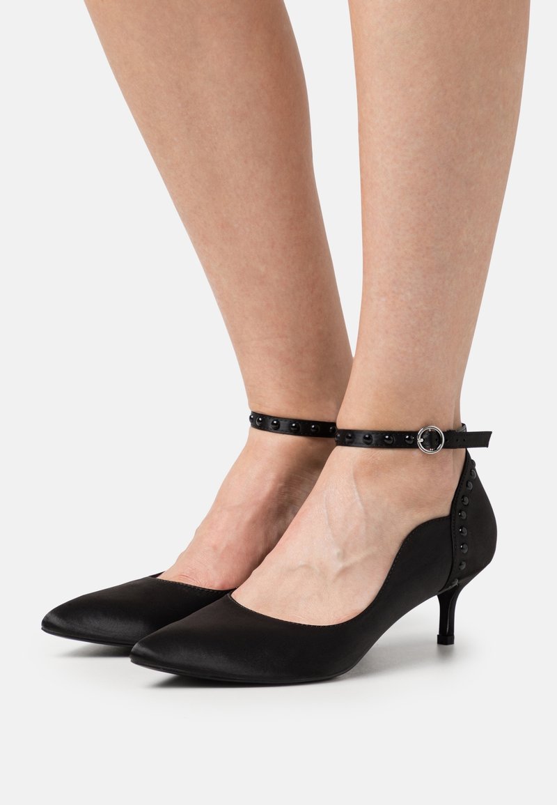 Escarpins noirs à bout pointu et talons kitten avec brides chevilles cloutées et fermetures à boucle portés pieds nus sur un fond blanc uni.