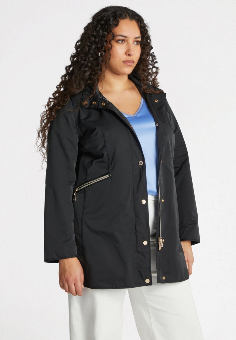 Zwarte waterdichte jas met een rits en drukknopen, voorzien van gouden accenten, zijzakken en een capuchon, gestyled met een blauwe top.