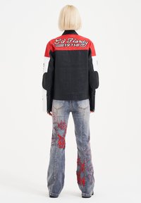 Black leather jacket with red and white accents, featuring embroidered text. Worn with light blue denim jeans, adorned with red patterns.