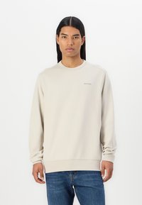 Felpa beige chiaro con maniche lunghe, scollo rotondo e discreto dettaglio del logo sul petto. Abbinata a jeans blu.