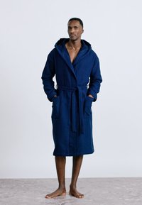 Άνδρας όρθιος με γυμνά πόδια, φοράει σκούρο μπλε μπάνιο με κουκούλα μέχρι τα γόνατα, με δεμένη ζώνη και τα χέρια στις τσέπες.