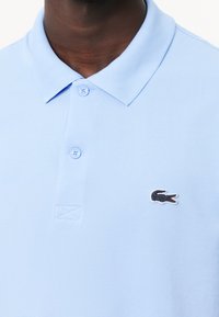 Homme portant un polo bleu clair avec deux boutons fermés et un petit logo crocodile noir sur la poitrine gauche.
