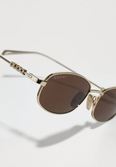 Gafas de sol metálicas doradas con lentes marrones; presentan un marco ovalado, bisagras laterales y la marca "GUCCI" en ambos lentes y patillas.