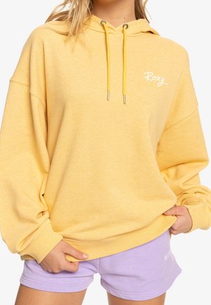 Osoba w za dużej żółtej bluzie z kapturem ze sznurkami i małym białym logo „Roxy”, nosząca jasnofioletowe szorty.