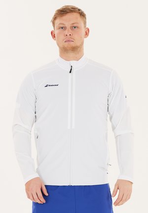 Ung mand med kort blondt hår iført en hvid Babolat sportstrøje med lynlås og blå shorts, stående foran en ensfarvet baggrund.