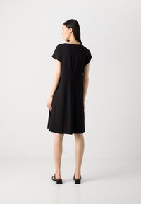 Black short-sleeved dress with a relaxed fit, rounded neckline, and smooth texture, falling just above the knees. Paired with block-heeled sandals.