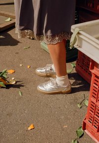 Sneakers metallici argento con un design texturizzato, suola di colore chiaro, abbinati a calze bianche a coste, accanto a bucce d'arancia sparse e casse.