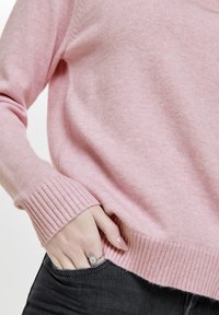 Rosa Strickpullover mit gerippten Bündchen und Saum. Weicher, strukturierten Stoff. Hand ruht in der Tasche der schwarzen Jeans, ein silberner Ring ist sichtbar.
