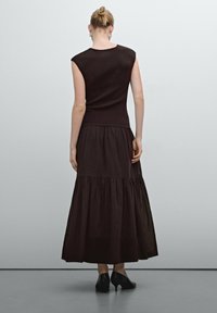 Tummanruskea, hihaton mekko, jossa on istuva yläosa ja kerroksellinen, laineileva helma. Valmistettu kevyestä kankaasta, siinä on hienovarainen tekstuuri ja yksinkertainen muotoilu.