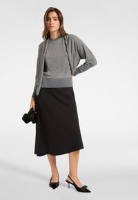 Cárdigan de punto gris sobre una camiseta gris, combinado con una falda midi negra y unos tacones de punta negra con detalles de lazo, sosteniendo un bolso negro.