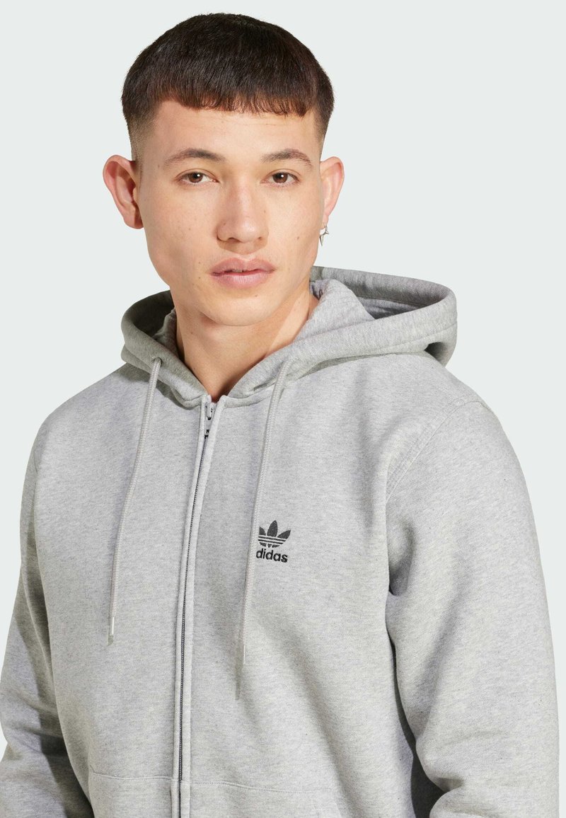 Sudadera con cremallera gris hecha de una mezcla de algodón. Presenta cordones, bolsillos tipo canguro y un pequeño logo negro en el lado izquierdo del pecho. Textura suave.