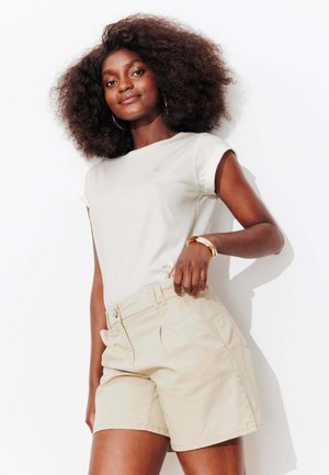 Femme aux cheveux bouclés et volumineux, portant un haut beige à manches courtes et un short beige, debout avec la main posée sur la taille, sur un fond blanc.