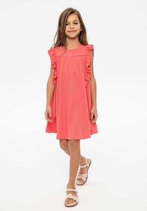 Robe corail avec des manches flottantes et une texture douce, évasée à la taille. Associée à des sandales claires avec un détail en nœud.