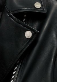 Col de veste en cuir noir avec boutons-pression argentés et une fermeture éclair argentée partiellement visible sur un fond sombre.