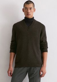 Dunkelgrüner V-Ausschnitt-Pullover aus strukturiertem Strickstoff, mit gerippten Bündchen und Saum. Trägt man über einem schwarzen Rollkragenpullover.
