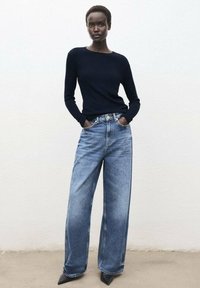 Marineblauer Pullover kombiniert mit hochgeschnittenen, weit geschnittenen hellblauen Jeans und spitzen schwarzen Pumps, vor einem schlichten hellen Hintergrund.