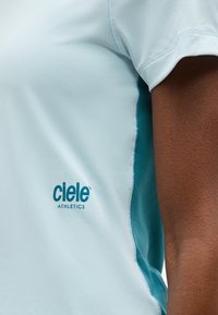 Κοντινή λήψη αθλητικής μπλούζας ανοιχτού μπλε χρώματος με πλευρικό πάνελ και μανίκι σε τιρκουάζ απόχρωση, που φοριέται από ένα άτομο με σκούρο δέρμα, παρουσιάζοντας το λογότυπο "ciele ATHLETICS" στο στήθος.
