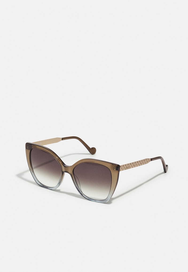 Overdimensjonerte solbriller med en gradient brun tone, transparent brun ramme og teksturerte brune vevde detaljer på stengene.
