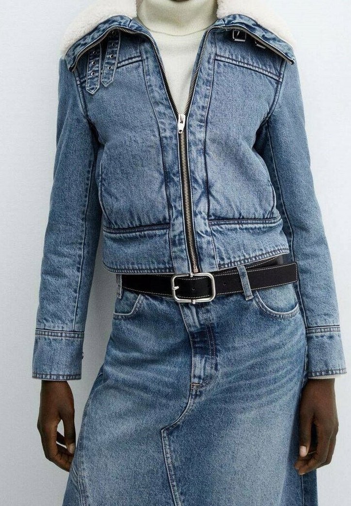Personne portant une veste en jean bleu clair avec une fermeture éclair, une ceinture noire et une jupe en jean assortie par-dessus un pull à col roulé blanc.