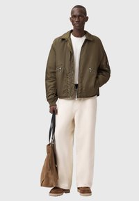 Olivengrøn bomberjakke med to forlommer med lynlås, båret over en hvid skjorte og lysebeige bukser med vide ben, holder en brun tote-taske.