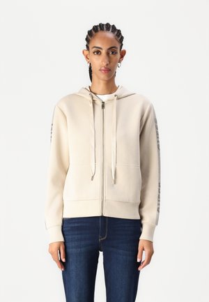 Sudadera cremosa con cremallera, capucha ajustable, bolsillos frontales y detalles impresos a lo largo de las mangas. Combinada con unos vaqueros azul oscuro.