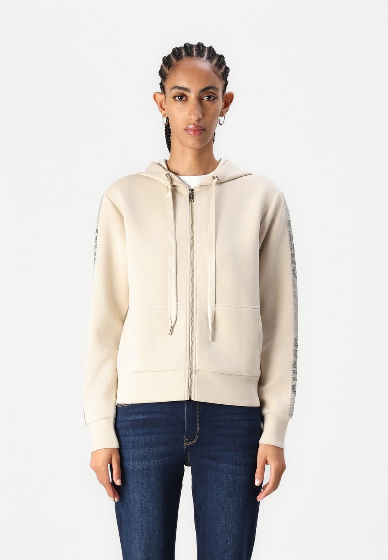 Sudadera cremosa con cremallera, capucha ajustable, bolsillos frontales y detalles impresos a lo largo de las mangas. Combinada con unos vaqueros azul oscuro.