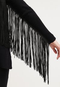 Höger arm sträckt ut, klädd i en svart långärmad plagg med långa svarta fransar hängande från ärmsluten.