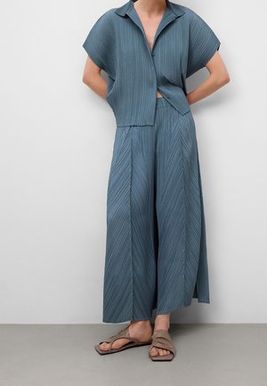 Femme portant un haut boutonné bleu plissé à manches courtes et un pantalon large, associé à des sandales marron clair, debout contre un mur uni.