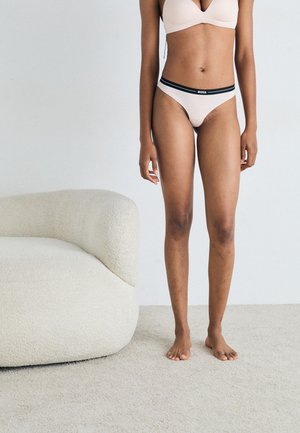 Soutien-gorge rose clair et string assorti avec une bande de logo noire ; tissu lisse ; modèle debout pieds nus près d'un canapé crème texturé.
