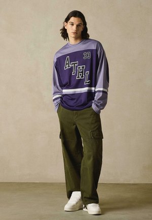 Jeune homme aux cheveux mi-longs portant un maillot de sport violet avec l'inscription "ATHL 33", un pantalon cargo olive et des baskets blanches, debout les mains dans les poches.