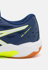 Férfi sportcipő sötétkék szintetikus felsőrésszel, fehér középtalppal, élénk sárga részletekkel és narancssárga gumitalppal. A "GEL-ROCKET 11" logó látható.