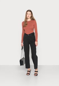 Camisola longa de manga ribeteada em rust emparelhada com jeans oversized de cintura alta pretos. Modelo segura uma mala preta texturizada e usa sandálias de tiras pretas.