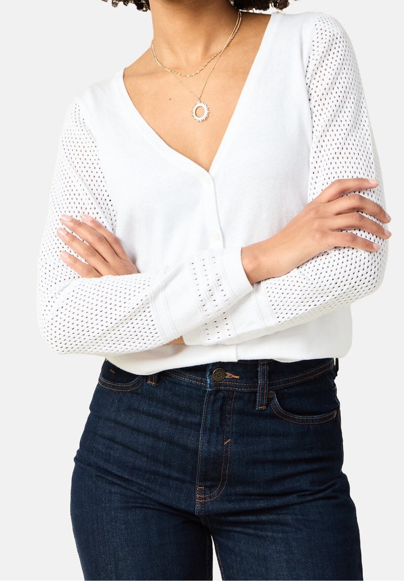 Cardigan blanc en maille avec un col en V, présentant des manches ajourées. Associé à un jean ajusté bleu foncé. Colliers dorés superposés en accessoires.