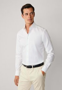 Uomo che indossa una camicia bianca aderente, pantaloni beige e una cintura nera, in piedi con una mano in tasca, mentre guarda di lato.