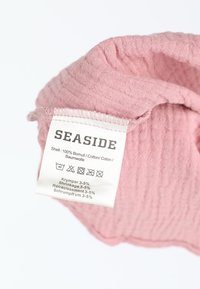 Lyserød bomuldsstof med en struktureret overflade, foldet over og viser en plejelabel. Etiketten indeholder vaskeanvisninger og mærkenavnet "SEASIDE."